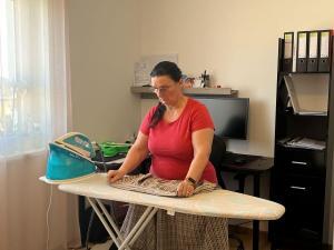 Woman in a pink shirt ironing a plaid shirt on an ironing board in a bright room with a window nearby.