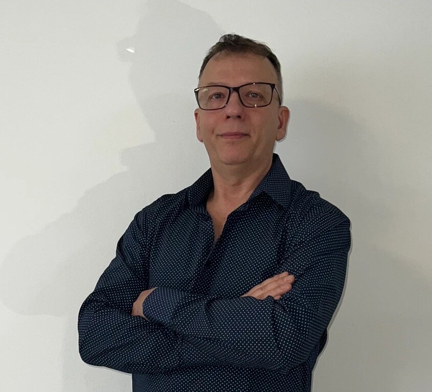 Man with Short Hair and Glasses Standing Against a Plain Light Wall Arms Crossed Wearing a Dark Blue Dotted Shirt